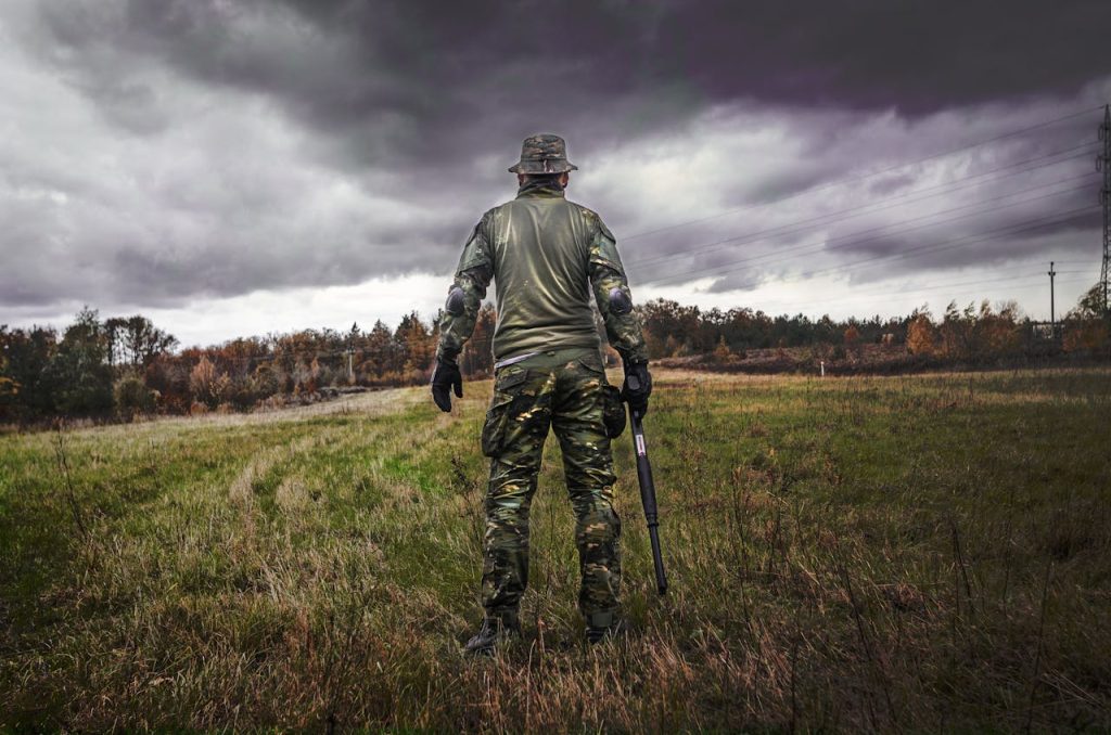 man-in-camouflage-suit-holding-shotgun-669277 A soldier in camouflage uniform with a rifle stands in a grassy field under a cloudy sky.